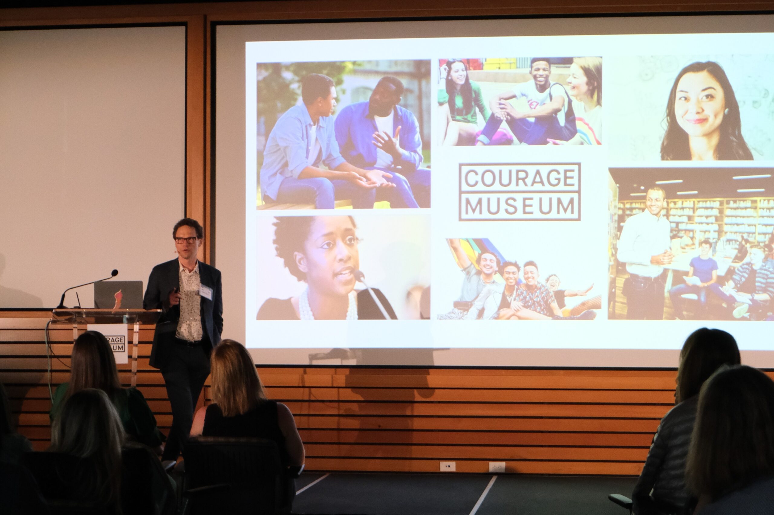 Jake Barton presenting on stage during an event at the Courage Museum with a presentation displayed on a large screen.